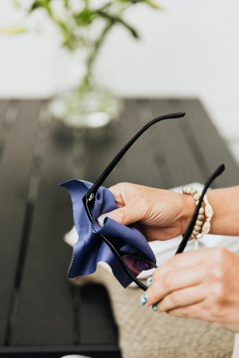 A person cleaning prescription glasses with a soft blue microfibre cloth, demonstrating how to clean anti-reflective lenses without scratching them.