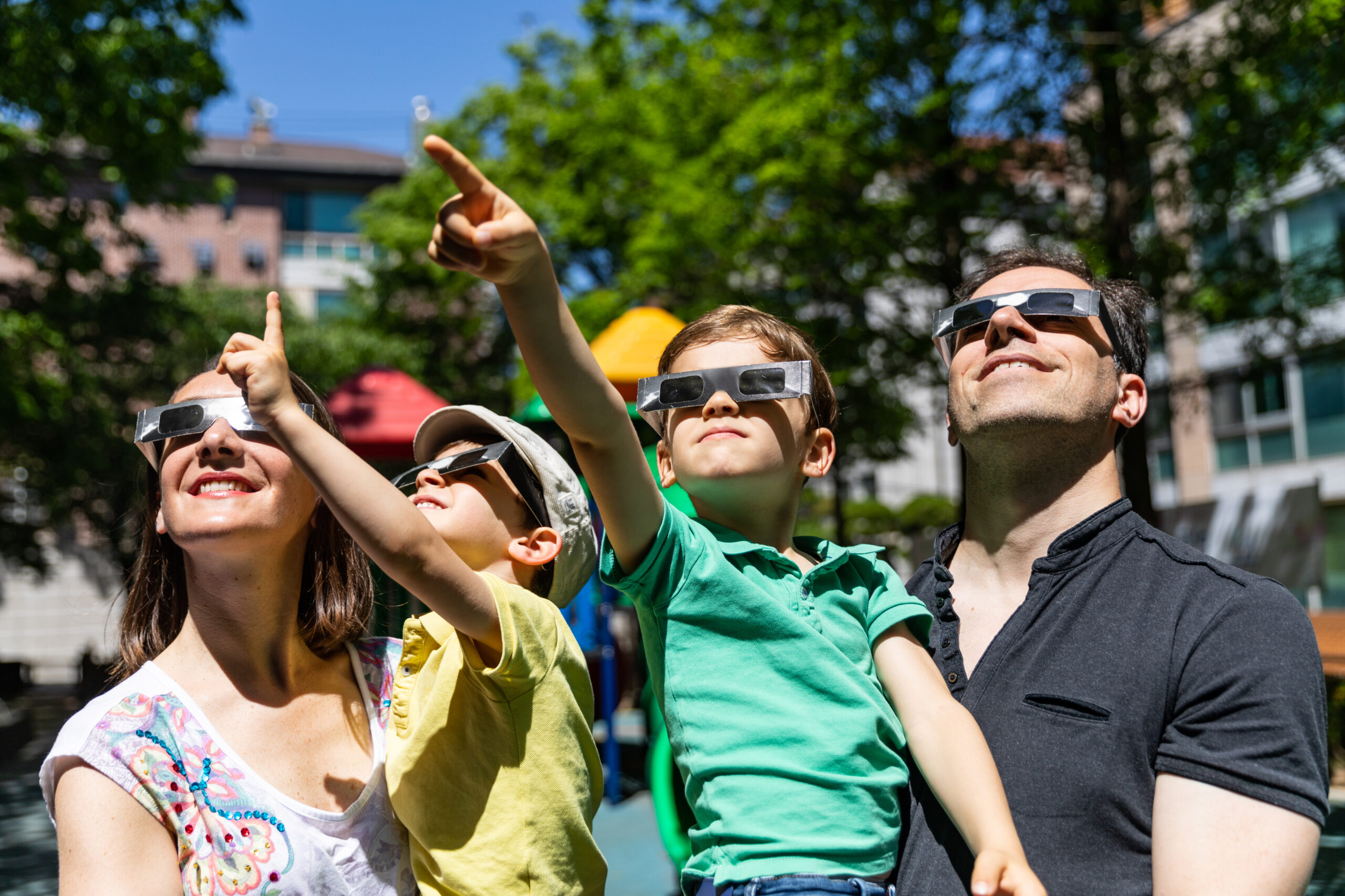 Family of four wearing solar eclipse glasses and pointing at the sky in a sunny park